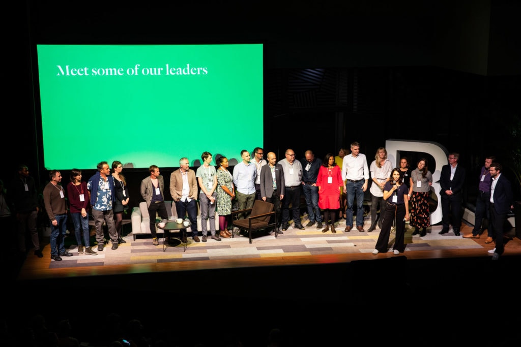 A group of people from B Corporation stand in front of a green screen displaying the text 'Meet some of our Leaders'