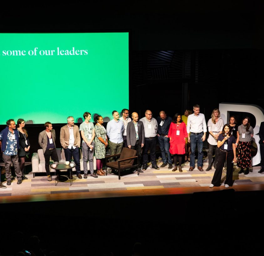 A group of people from B Corporation stand in front of a green screen displaying the text 'Meet some of our Leaders'