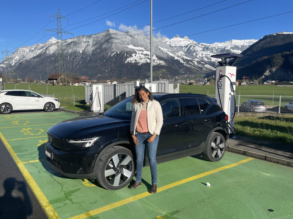 Vineeta standuing next to a black electric Volvo plugged into a charger with snowy mountains in the background